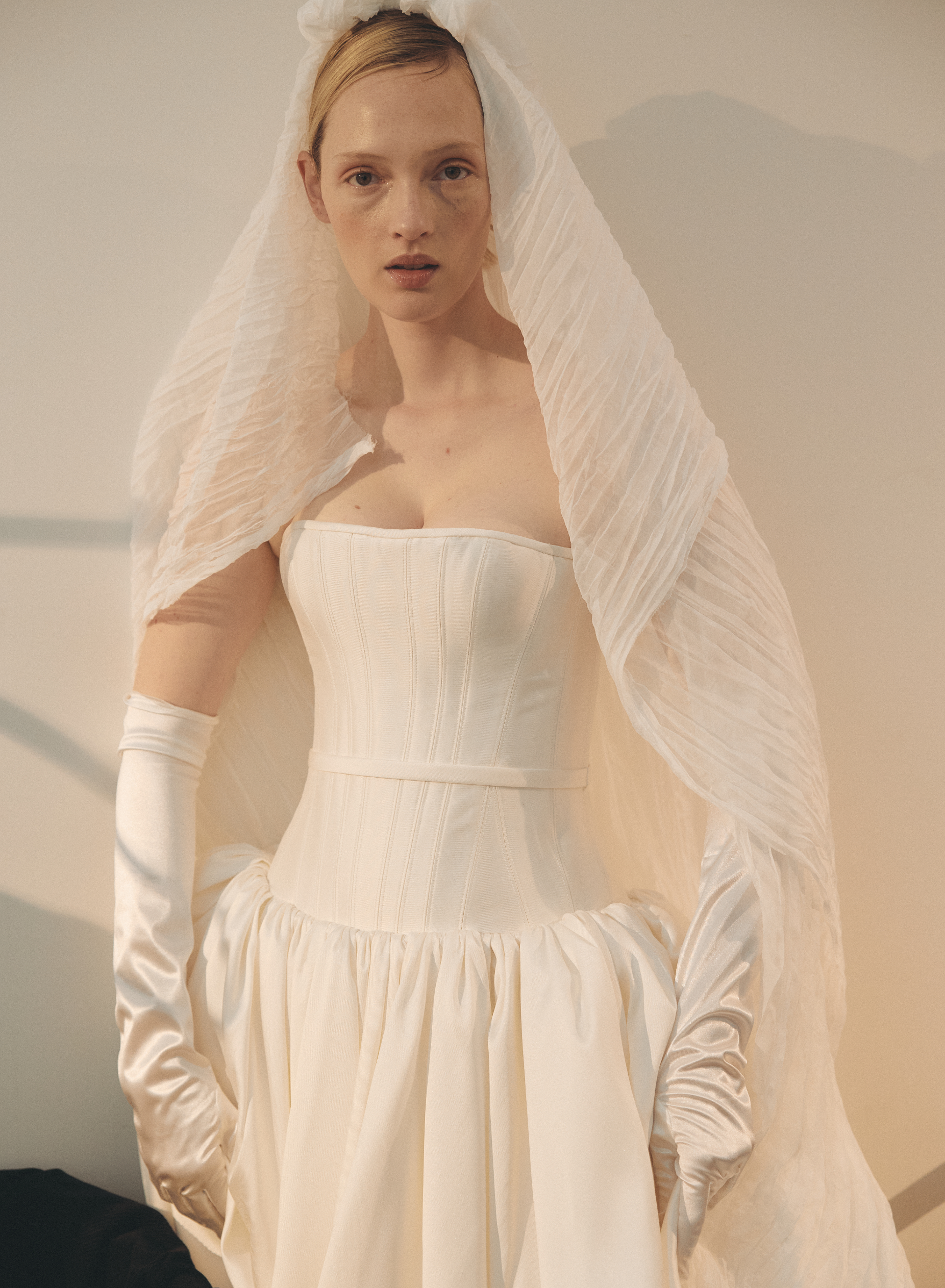 Woman wearing a white wedding dress with a veil and gloves indoors.